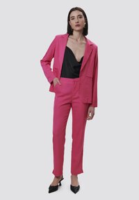 Felroze blazer en bijpassende broek, gladde textuur, nauwsluitend ontwerp, gecombineerd met een zwarte satijnen top en zwarte puntige hakken.