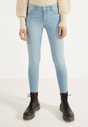 Kobieta w jasnoniebieskich obcisłych dżinsach z niewielkimi przetarciami i czarnych sznurowanych butach wojskowych, stojąca na jednolitym tle.