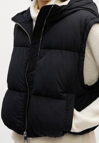 Veste sans manches matelassée noire avec fermeture éclair à l'avant, portée sur un pull blanc en tricot à manches longues.