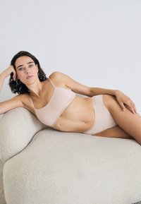 Vrouw met kort donker haar, gekleed in huidkleurig ondergoed, liggend op een textuurrijke crèmekleurige bank tegen een witte muur.