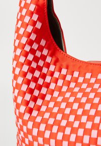 Rød veske med et vevd design som har alternerende rosa firkanter. Jevn tekstur med en blank finish, rund form og sydd kanter.
