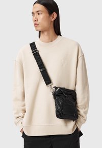 Giovane con capelli lunghi e scuri indossa una felpa beige e incrocia una borsa a spalla nera imbottita con una larga tracolla sul petto.