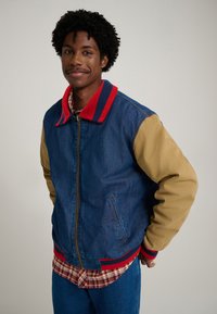 Denim bomber jacket featuring a blue body, khaki sleeves, and a red and navy striped collar. Paired with a plaid shirt and blue jeans.