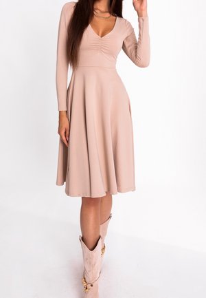 Kobieta w beżowej sukience za kolano z długim rękawem i marszczonym dekoltem oraz jasnoróżowymi kowbojkami, stojąca na białym tle.