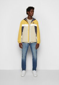 Giacca a vento colorata con pannelli gialli, beige e blu navy; presenta una chiusura con zip, cappuccio regolabile e due tasche anteriori. Indossata con jeans azzurri.