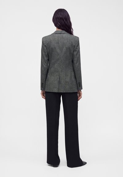 Femme aux longs cheveux foncés avec un blazer gris à carreaux et un pantalon noir à jambe large, debout de dos sur un fond uni.