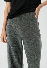 Glitzernde schwarze Hose mit strukturiertem Finish, weitem Beinansatz und elastischem Bund. Glattes Material mit subtilen Glanzeffekten.