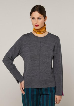 Femme aux cheveux foncés portant un pull gris à manches longues, une écharpe jaune moutarde à pois autour du cou, et un pantalon bleu sarcelle à rayures verticales.