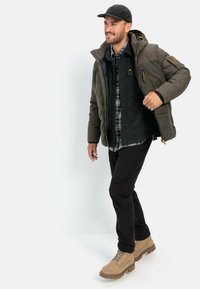 Polstret olivenfarvet jakke med hætte, lagdelt over en mørk flannelskjorte. Sorte bukser og brune støvler med strukturerede såler.