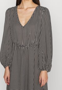 Svart och vit houndstooth-mönstrad klänning med djup V-ringning, puffärmar och elastisk midja för en flödande silhuett.