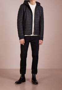 Svart vadderad jacka med huva, med dragkedjestängning och ribbade ärmslut, bärs över en krämfärgad kabelstickad tröja och svarta jeans. Svarta stövlar.