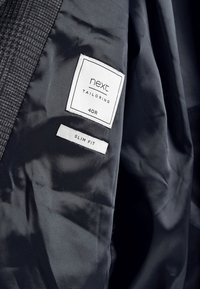 Schwarzes maßgeschneidertes Jackeninnenfutter mit strukturiertem Stoff. Etiketten mit der Aufschrift "next tailoring" und "40R" auf weißen Etiketten; "Slim Fit" in kleinerem weißem Text.