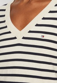 Pull à col en V rayé en crème et marine, avec une texture en maille fine et un petit détail de logo. Points forts : un design classique avec des rayures horizontales.