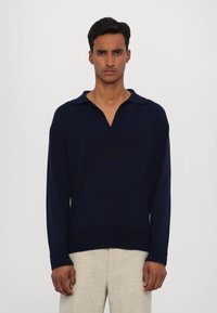 Námořnický modrý pletený pulovr s výstřihem do V a límečkem, dlouhými rukávy a rovním lemem, spárovaný se světlými strukturovanými kalhotami.