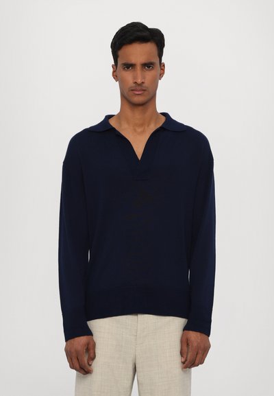 Pull-over en tricot bleu marine avec un col en V et un col, doté de manches longues et d'une coupe droite, associé à un pantalon texturé de couleur claire.