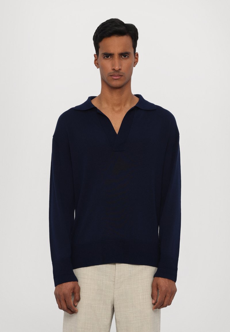 Pullover lavorato a maglia blu navy con scollo a V e colletto, dotato di maniche lunghe e orlo dritto, abbinato a pantaloni chiari e strutturati.