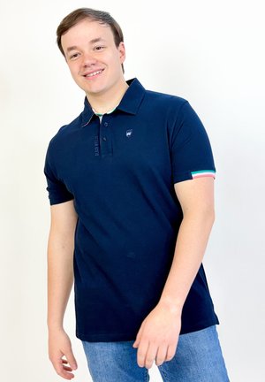 Jeune homme souriant, portant un polo bleu marine avec des bordures rayées aux manches et un jean bleu clair, debout sur un fond blanc.