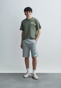 Μπλούζα από βαμβάκι σε ελιά πράσινο χρώμα, με κοντά μανίκια και στρογγυλή λαιμόκοψη, συνδυασμένη με γκρι αθλητικές βερμούδες με μπλε λογότυπο, λευκά αθλητικά παπούτσια και λευκές κάλτσες.