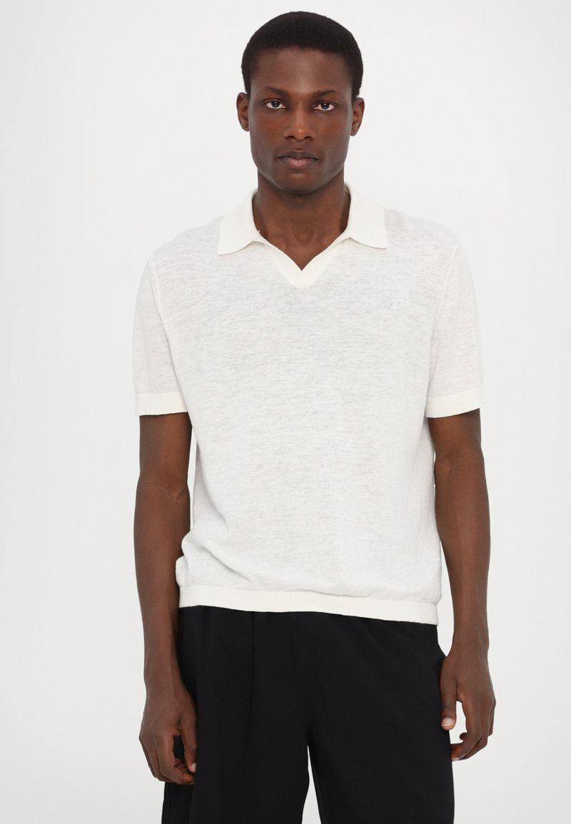 Polo bianco a maglia con scollo a V e maniche corte. Presenta un colletto a contrasto e orlo a coste, esibito su un modello contro uno sfondo neutro.