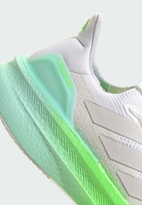 Weiße Sportschuh mit strukturiertem Obermaterial und hellgrünen Akzenten, markanten drei Streifen und einer gradierten grünen Außensohle, die mit "ULTRABOOST" beschriftet ist.
