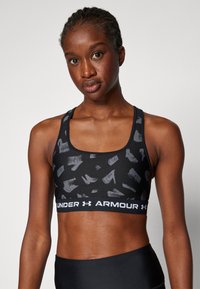 Melna sporta krūšturis ar teksturētu pelēku otas trieciena rakstu. Iezīmē plata jostas lente ar "UNDER ARMOUR" logotipu. Sacīkšu aizmugure.