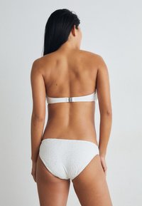 Bela bikinka z bandeau vrhom in teksturiranim visokim dnom, ki vsebuje krožne vzorce. Gladek material, minimalni detajli iz kovine.