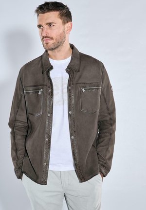 Braune Lederjacke mit zwei Brusttaschen, Druckknöpfen und kontrastierendem Nähen; getragen über einem weißen T-Shirt und grauen Hosen.