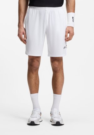 Mann trägt weiße Sportshorts, weiße Socken, weiße Turnschuhe, schwarzes Hemd und ein weißes Armband mit dem "BOSS"-Logo auf dem Armband und den Shorts.