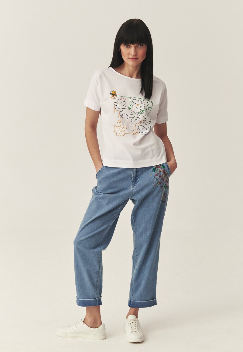 Frau trägt weißes T-Shirt mit Blumen- und Bienenmuster, lockere hellblaue Jeans mit Blumenstickerei auf der Tasche und weiße Turnschuhe.