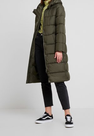 Personne portant un long manteau matelassé vert olive, un pull vert clair, un jean court noir et des baskets noir et blanc, debout devant un fond uni.