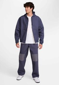 Mornarsko modra jopa s kapuco in vrvicami, sprednjimi žepi, kombinirana z ujemajočimi hlačami s teksturiranimi belimi pikčastimi našitki na sprednji strani. Bele superge.