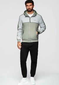 Letvægts, tofarvet jakke i grøn og grå, med hætte, lynlåslukning foran og sidelommer, parret med sorte bukser og hvide sneakers.