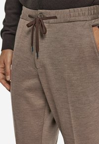 Brązowe dzianinowe spodnie mają elastyczny pas ze sznurkiem i zapięciem na guzik, kieszeniami po bokach oraz teksturowanym materiałem dla wygody.