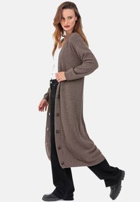 Cardigan long marron en matière douce, avec une coupe décontractée, des boutons sur le côté et des poignets côtelés, associé à un pantalon large noir et des chaussures.