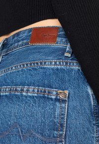 Jeans de ganga azuis com um patch de couro rotulado "Pepe Jeans London", com costura dourada e um design ondulado no bolso traseiro.
