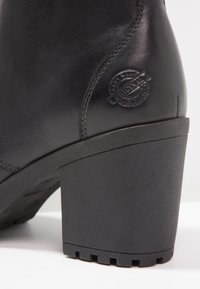 Botín de cuero negro hasta el tobillo con una superficie texturizada, puntera redonda y un tacón grueso. Presenta un emblema elevado en el lateral.