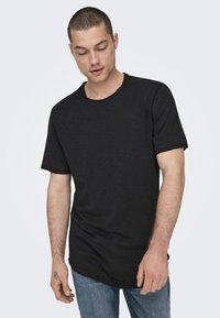 Schwarzes Kurzarm-T-Shirt aus weichem Stoff mit rundem Ausschnitt und rohen Kanten. Getragen mit blauen Jeans.