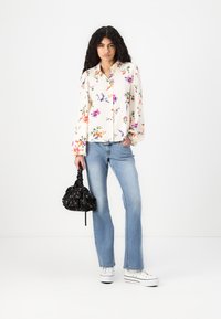 Floral patterned blouse in off-white fabric with long sleeves, paired with light blue jeans and a black drawstring bag with floral accents.