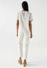 Femme aux longs cheveux bruns portant un top beige à manches courtes, un pantalon blanc et des sandales à talons marron, se tenant devant un fond blanc uni.