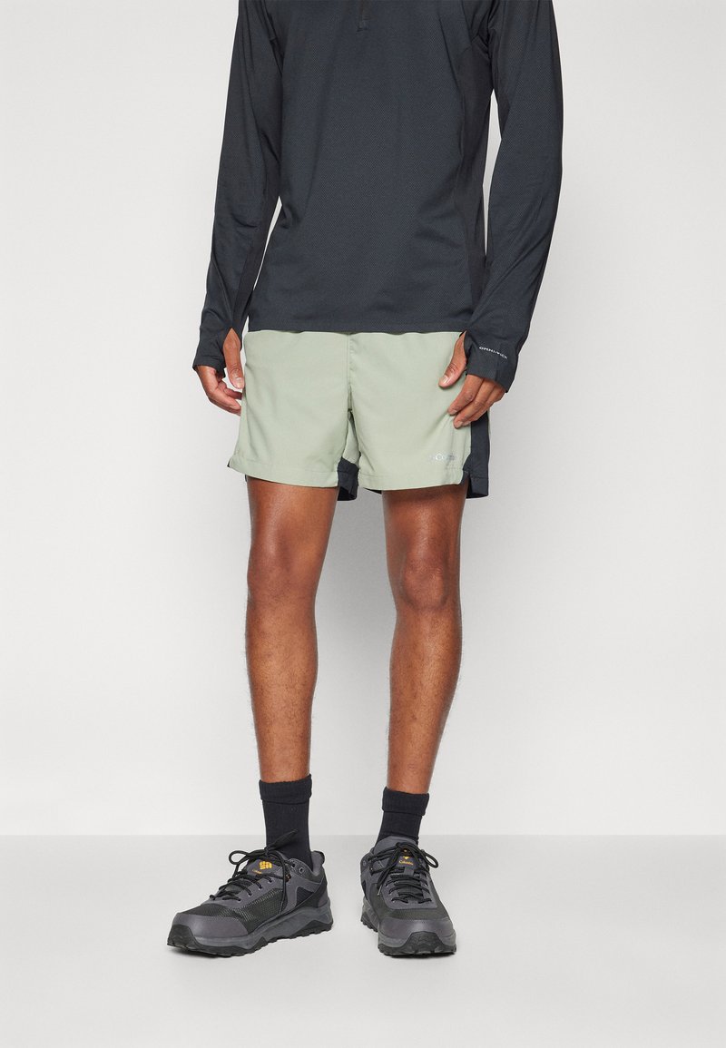 Pantalones cortos verde claro con aberturas laterales, combinados con una camiseta negra de manga larga. Zapatillas de senderismo grises y calcetines negros completan el atuendo.