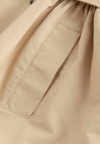 Tela beige con una textura suave, que presenta un pequeño bolsillo con un detalle de costura limpio, ligeramente recogido en la parte superior para agregar volumen.