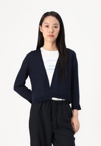 Navy blue cropped cardigan with long sleeves, V-neck design, ribbed hem, worn over a white graphic T-shirt and black trousers.