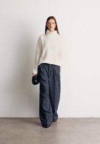 Suéter oversized branco de gola alta combinado com calças largas de padrões escuros. Modelo segura uma pequena bolsa preta. Fundo liso.