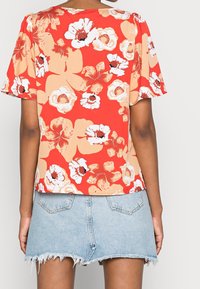 Blusa corta de manga corta con flores rojas, crema y naranjas, confeccionada en tejido ligero. Llevada con una falda de jean deshilachada, mostrando la vista trasera.