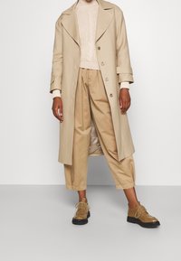 Cappotto trench doppio petto beige sopra un maglione in maglia a treccia crema e pantaloni beige a pieghe, abbinati a scarpe in pelle beige con lacci e suole nere.