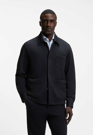 Homme portant une veste sombre boutonnée avec trois poches devant par-dessus une chemise bleu clair à col, debout devant un fond blanc uni.