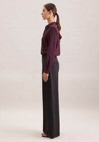 Bordeauxrotes Langarmhemd mit Kragen, kombiniert mit schwarzen, hochbundigen Hosen. Die Hosen haben ein gerades, weites Bein und dezente Taschendetails.