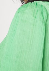 Tessuto verde testurizzato con un aspetto spiegazzato. Presenta un collo arricciato e una silhouette fluttuante, creando una vestibilità rilassata.