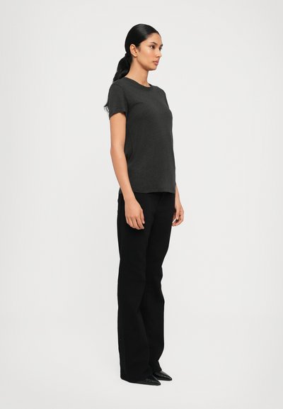 Mujer con cabello largo y oscuro recogido en una coleta, vestida con una camiseta negra de manga corta, pantalones negros y zapatos negros puntiagudos, de pie mirando hacia la derecha.