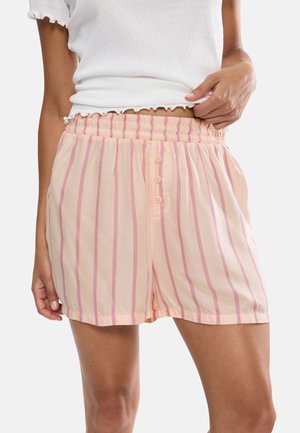 Kobieta ubrana w brzoskwiniowe szorty z pionowymi różowymi i niebieskimi paskami oraz trzema guzikami na elastycznym pasku, w połączeniu z białym, krótkim topem.